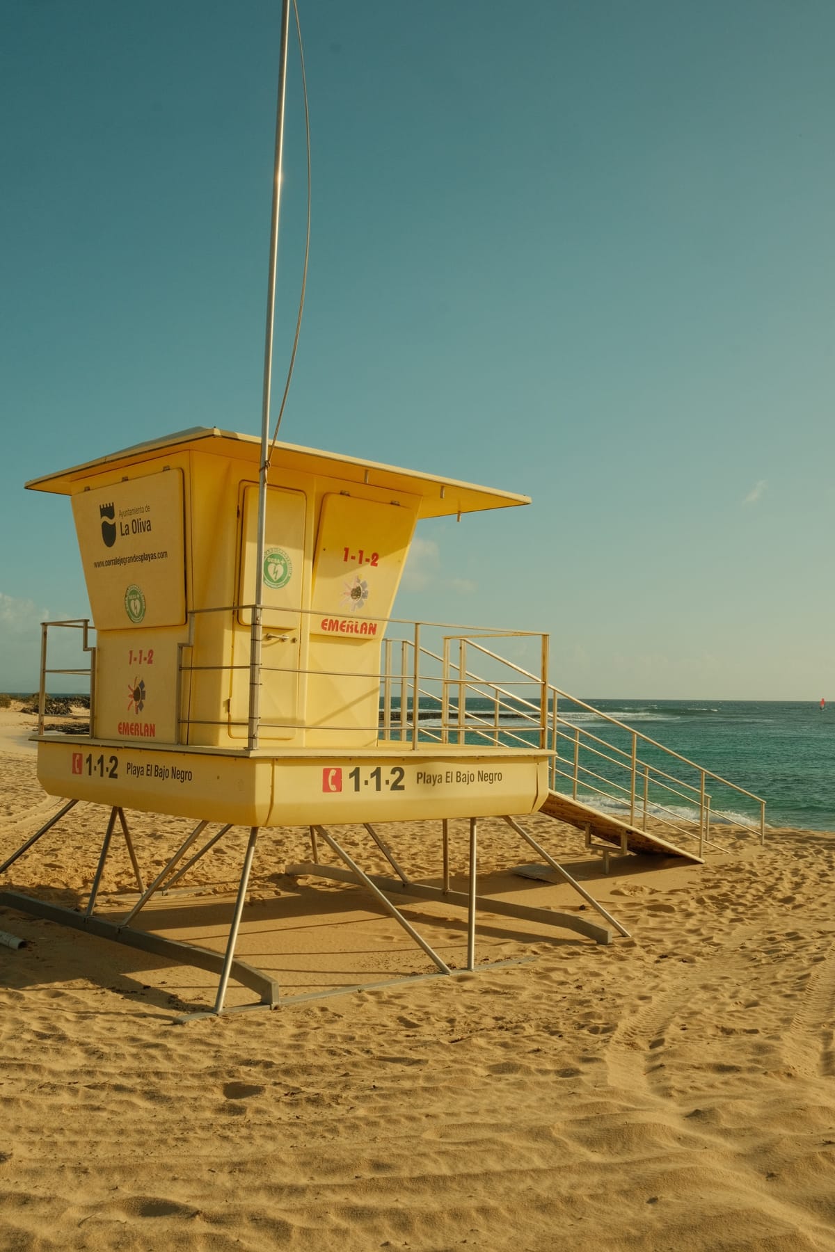 Ein gelber Rettungsturm auf Stelzen im Sand, dahinter das türkise Meer.